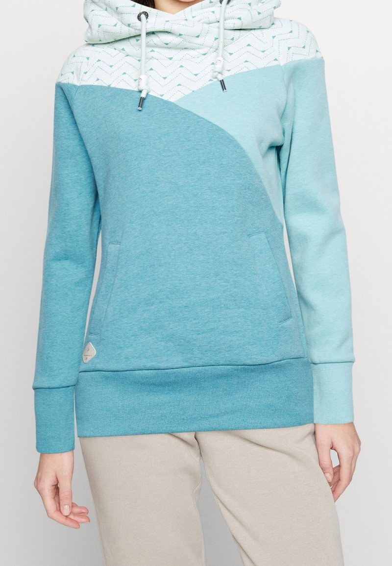 Femme portant un sweat à capuche bleu et blanc avec une poche avant et un pantalon beige, debout devant un fond clair uni.