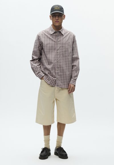 Camisa de manga larga con cuadros en marrón y beige, combinada con pantalones cortos beige hasta la rodilla. Zapatos negros y gorra negra con texto amarillo.