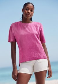 Short-sleeve, textured top in vibrant pink, paired with light-colored shorts. Smooth fabric with a relaxed fit. Suitable for casual wear.