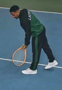 Costum sportiv verde și negru cu brandul Lacoste, ținând o rachetă de tenis portocalie, purtând adidași albi pe un teren de tenis interior albastru.