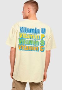 Kremfarget oversized t-skjorte som har fet, flerfarget tekst: "Vitamin U", "Vitamin P", "Vitamin S" og "Vitamin C" ordnet vertikalt.
