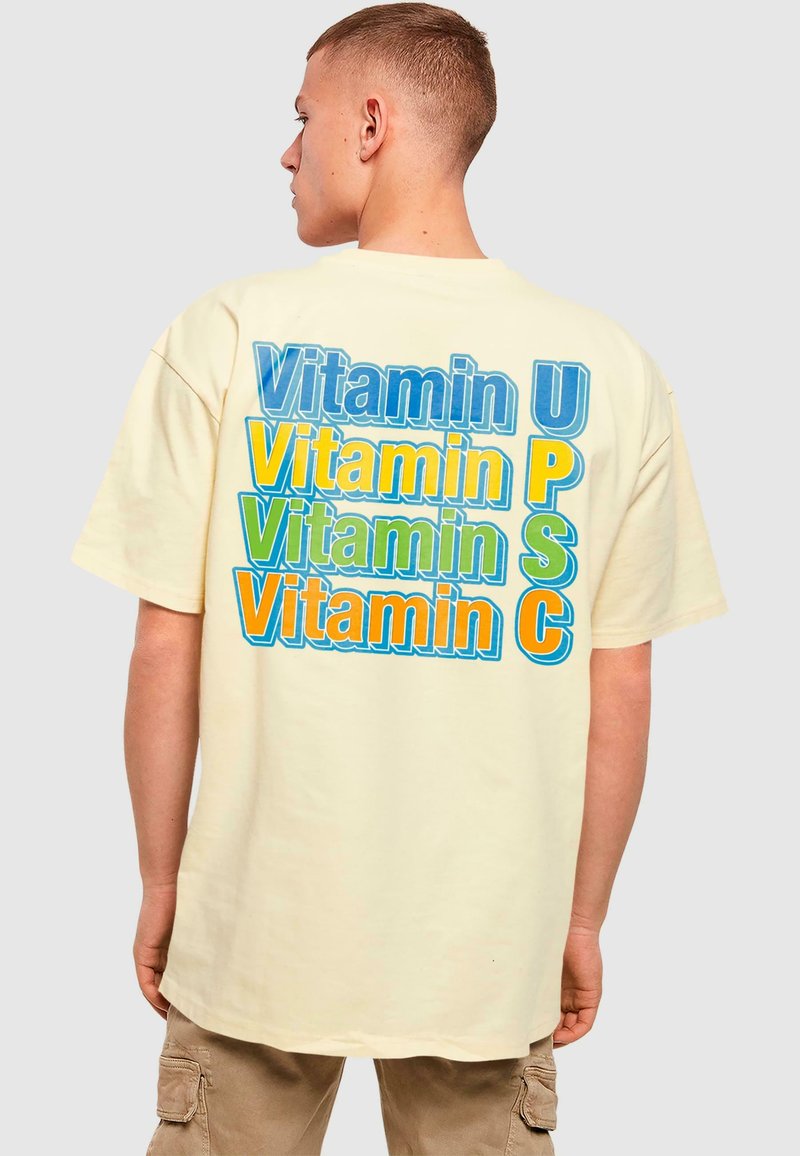 Kremfarget oversized t-skjorte som har fet, flerfarget tekst: "Vitamin U", "Vitamin P", "Vitamin S" og "Vitamin C" ordnet vertikalt.