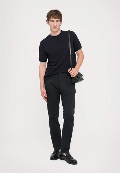 T-shirt bleu marine à manches courtes avec des bordures bleu clair, pantalons noirs et chaussures en cuir noires. Le modèle tient un petit sac à bandoulière noir.