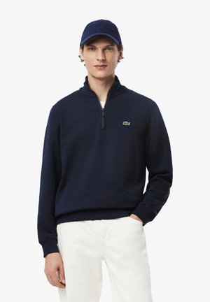 Navy blue zip-up sweatshirt made of cotton blend, featuring a ribbed collar and cuffs, with a small green logo on the chest. Model wears white pants.