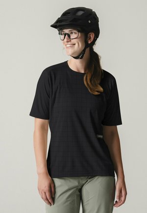 Frau mit schwarzem Fahrradhelm, Brille, schwarzem Kurzarmshirt und hellgrüner Hose, die lächelt und zur Seite schaut.