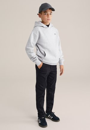 Hallide hall kapuutsiga, millel on eesmine tasku, mustad püksid ja tume jalatsid valgete triipudega. Kandes halli mütsi, seisab käed taskutes.