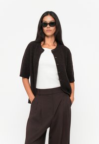 Dark brown short-sleeve cardigan with a round neckline and button closure, worn over a white top, paired with high-waisted brown trousers.