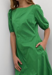 Grön klänning med puffärmar, rundad halsringning, sidofickor och en mjuk, glänsande yta. Enkel design utan mönster.