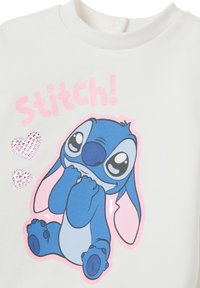 Witte sweatshirt met een blauwe cartoonfiguur met grote oren en expressieve ogen. "Stitch!" tekst in glinsterend roze, met hartaccenten.