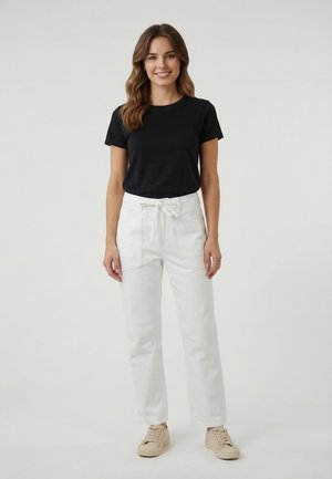 Femme debout et souriante, portant une chemise noire à manches courtes, un pantalon taille haute blanc avec ceinture nouée, et des baskets beige.