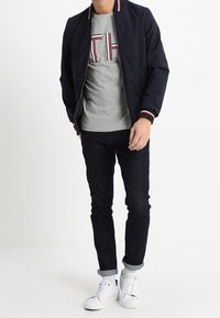 Veste de pilote de marine avec des accents rouges et blancs, t-shirt gris avec logo, jeans foncés, baskets blanches et manches retroussées.
