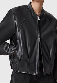 Giacca bomber nera in pelle con un design aderente, polsini a coste e colletto alto con zip frontale. Tessuto liscio e dettagli minimali.