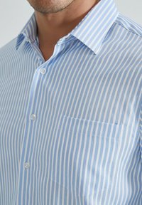 Chemise à col bleu clair et blanc rayée avec un tissu texturé, comportant une poche poitrine gauche et des boutons blancs.