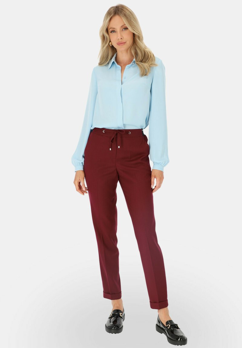 Blondynka w jasnoniebieskiej bluzce z długim rękawem, bordowych spodniach z troczkami i czarnych mokasynach, stoi na białym tle.