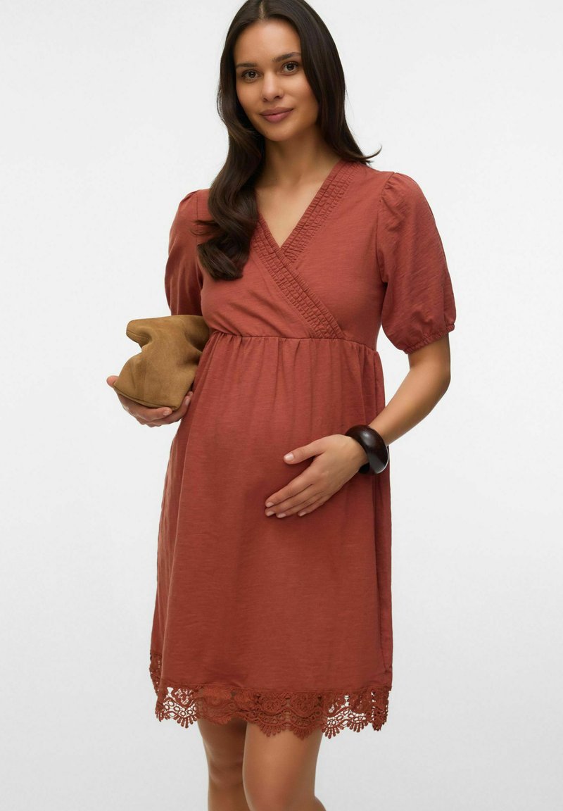 Femme enceinte aux longs cheveux foncés portant une robe portefeuille couleur rouille avec une bordure en dentelle, tenant une pochette beige et posant une main sur son ventre.