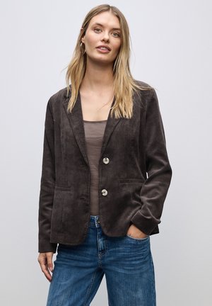 Blazer en velours côtelé marron avec revers cranté, deux boutons et poches latérales. Porté par-dessus un haut beige ajusté et associé à un jean bleu.
