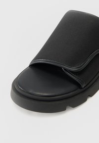 Black slide sandal featuring a smooth synthetic upper and a thick rubber sole with a textured finish. Open toe design with a wide strap.