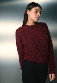 Bordeauxrød, blød sweater med rund hals og lange ærmer, der har en struktureret overflade. Kombineret med sorte, tætsiddende bukser.