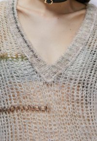 Zbliżenie na osobę noszącą jasny, luźno dziergany sweter v-neck na nagiej klatce piersiowej z czarnym naszyjnikiem choker.
