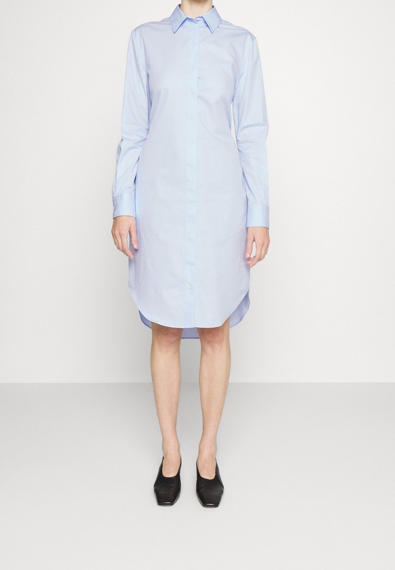Jasnoniebieska sukienka koszulowa z długim rękawem, zakończona szpiczastym kołnierzykiem, z zapięciem na guziki i zaokrąglonym dołem; w połączeniu z czarnymi butami. Gładka tekstura materiału.