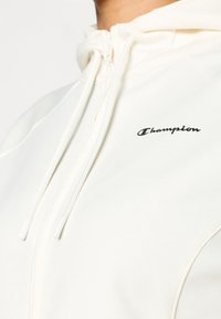 Μπεζ φούτερ με κουκούλα από απαλό ύφασμα, με φερμουάρ, κορδόνια και μικρό μαύρο λογότυπο "Champion" στην αριστερή πλευρά.