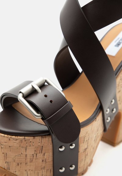 Πέδιλο με ψηλό τακούνι και πάτο από φελλό, σκουρόκαφέ δερμάτινα λουριά, ασημένια αγκράφα και μεταλλικά καρφιά στο πίσω μέρος του τακουνιού.