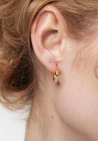 Złota kolczyk w kształcie obręczy, z fakturowaną powierzchnią i błyszczącym akcentem, noszony w przekłutym uchu, na jasnym tle.