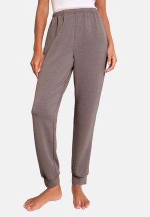Vrouw draagt taupekleurige joggingbroek met elastische taille en omgeslagen enkels, staat blootsvoets tegen een effen achtergrond.