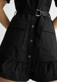 Abito nero con fondo svasato. Presenta due tasche frontali, chiusura con bottoni e una cintura abbinata con fibbia argentata. Tessuto dalla consistenza liscia.