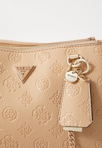 Borsa a tracolla beige con texture, motivi in rilievo, logo triangolare dorato e un charm con un'etichetta rotonda e una catena decorativa. Chiusura con cerniera.