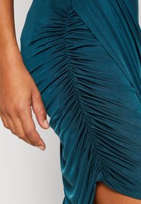 Gros plan d'un tissu de robe turquoise froncée avec une couture rassemblée et la main gauche d'une personne reposant à côté.