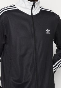 Svart Adidas-jacka med vit dragkedja och krage, med tre vita streck längs axlarna och en liten logotyp på bröstet.