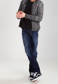Giacca in pelle grigia con dettagli trapuntati sulle spalle, t-shirt nera, jeans blu scuro e scarpe da ginnastica nere e bianche. Posizione in piedi con le mani unite.
