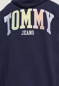 Navy tröja med huva som har en stor "TOMMY" logotyp i flerfärgade broderade bokstäver och "JEANS" i vitt nedan. Mjuk textur, design med huva.