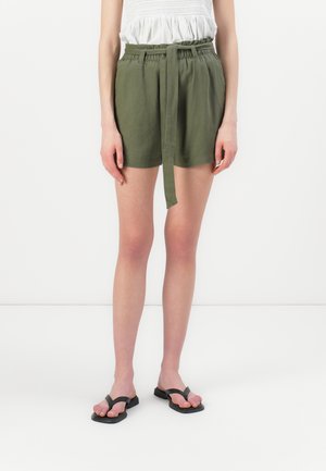 Femme portant un short taille haute vert olive avec une ceinture en tissu, un haut blanc sans manches, et des sandales noires à bout carré, debout.