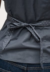 Γκρι υφασμάτινη ζώνη με λευκά πουά δεμένη σε φιόγκο γύρω από ένα ταιριαστό γκρι πουκάμισο, πάνω από μπλε τζιν με πορτοκαλί ραφές.