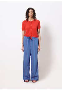 Roter strukturierter Kurzarm-Cardigan mit vorderem Band und blau gestreiften weit geschnittenen Hosen mit roten Streifen, kombiniert mit schwarzen Sandalen.