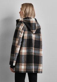 Cappotto a quadri di lana nei toni del marrone, nero e bianco, dotato di cappuccio, grandi motivi a quadri e polsini a coste. La vista posteriore mostra una vestibilità rilassata.