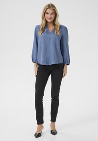 Blusa blu a maniche lunghe con scollo a V e polsini elasticizzati, abbinata a jeans skinny neri e mule nere a punta, su uno sfondo semplice.