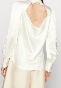 Donna che indossa una blusa di raso bianca con schiena scoperta che rivela un piccolo tatuaggio di testa di alieno sulla parte superiore della schiena.