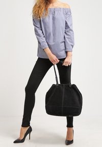 Černá semišová hobo taška s kulatým tvarem, vybavená dvojitými uchy a viditelnými švy. Modelka má na sobě pruhované tričko s odkrytými rameny.