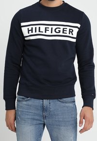 Marlová modrá mikina s kulatým výstřihem vyrobená z bavlněné směsi, s výrazným bílým logem "HILFIGER" přes hruď na horizontálním pruhu.