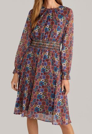 Kobieta w sukience do kolan z długimi rękawami, ozdobionej wzorem wielobarwnych kwiatów i z marszczonym pasem, stojąca na jednolitym tle.