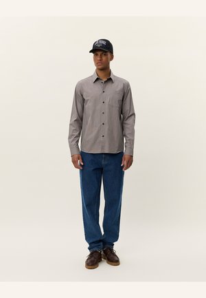 Heren geruite overhemdbloes in bruin en beige, gecombineerd met losse blauwe denim jeans en bruine veterschoenen. Zwarte pet met logo.
