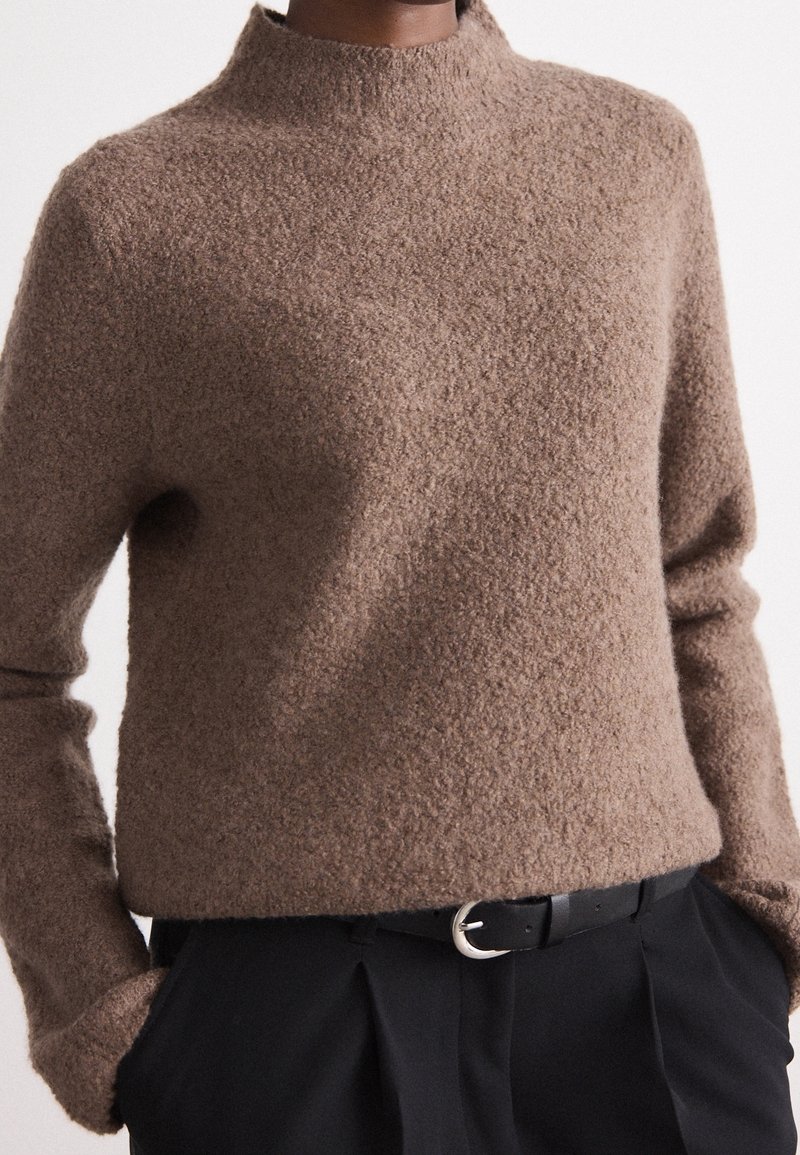 Pull marron texturé à manches longues porté avec un pantalon noir plissé et une ceinture noire avec une boucle argentée, les mains dans les poches.