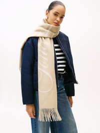 Écharpe en laine beige avec des franges, présentant un léger motif. Associée à une doudoune navy et une chemise rayée, portée par un modèle sur fond uni.