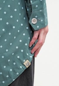 Zielona koszula w jasnoniebieskie grochy, z zapięciem na guzik przy mankiecie oraz metką z logo marki przy dolnym brzegu, wykonana z gładkiej, teksturowanej tkaniny.