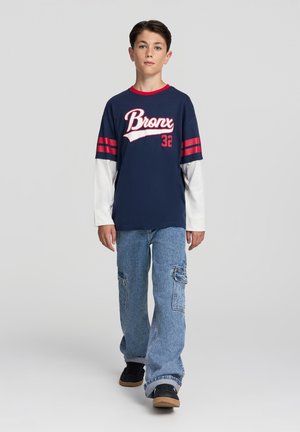 Maglietta blu navy con "Bronx 32" in testo bianco e rosso, maniche lunghe bianche, abbinata a jeans cargo blu chiaro e scarpe nere.