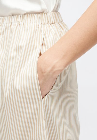 Ruka zasunutá do kapsy béžových a bílých pruhovaných kalhot s elastickým pasem na bílém pozadí.