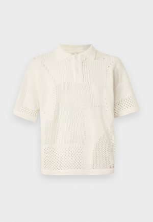 Mand iført solbriller, cremefarvet kortærmet strikket polo med netmønstre, løse mørke mønstrede vidde bukser, stående mod ensfarvet baggrund.